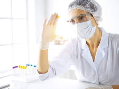 Koruyucu gözlüklü profesyonel kadın bilim adamı güneşli laboratuvarda reaktörlerle araştırma tüpü kullanıyor. Tıp ve araştırma.