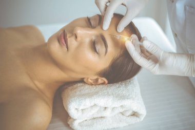 Güzellik uzmanı güneşli klinikte genç esmer kadının yüzüne şırıngayla güzellik prosedürü uyguluyor. Kozmetik ilaç ve ameliyat, güzellik iğneleri.