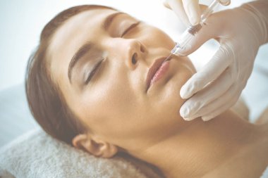 Güzellik uzmanı güneşli klinikte genç esmer kadının yüzüne şırıngayla güzellik prosedürü uyguluyor. Kozmetik ilaç ve ameliyat, güzellik iğneleri.