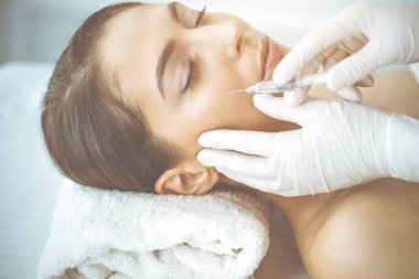 Güzellik uzmanı güneşli klinikte genç esmer kadının yüzüne şırıngayla güzellik prosedürü uyguluyor. Kozmetik ilaç ve ameliyat, güzellik iğneleri.