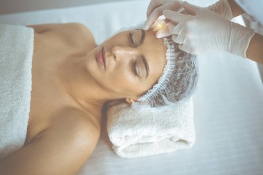 Güzellik uzmanı güneşli klinikte genç esmer kadının yüzüne şırıngayla güzellik prosedürü uyguluyor. Kozmetik ilaç ve ameliyat, güzellik iğneleri.