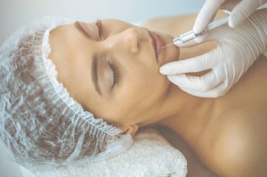 Güzellik uzmanı güneşli klinikte genç esmer kadının yüzüne şırıngayla güzellik prosedürü uyguluyor. Kozmetik ilaç ve ameliyat, güzellik iğneleri.