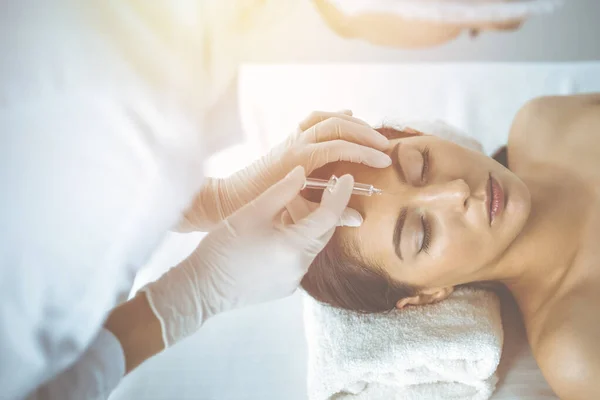 Beautician doing beauty procedure with syringe to face of young brunette woman in sunny clinic. Cosmetic medicine and surgery, beauty injections