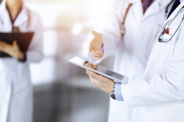 Bilinmeyen bir grup doktor, güneşli bir hastane ofisinde dururken bazı tıbbi isim kayıtlarını kontrol etmek için bir bilgisayar tableti kullanıyor. Doktorlar hastaları muayene etmeye ve yardım etmeye hazır. Tıbbi yardım, sigorta.