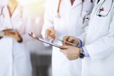 Tanınmayan doktorlar, iki erkek ve bir kadın, güneşli hastane ofisinde dururken tıbbi muayene sonuçlarını tartışıyorlar. Doktorlar ilaç kayıtlarını doldurmak için pano kullanıyorlar. Mükemmel sağlık.