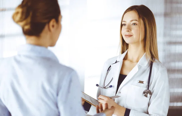 젊은 여성 의사와 환자들은 병원에서 함께 서 있는 동안 현재의 건강 검진에 대해 논의하고 있다. 여성 의사들은 클립보드를 사용하여 몇 가지 표시를 작성하고 있다. 완벽 함 — 스톡 사진