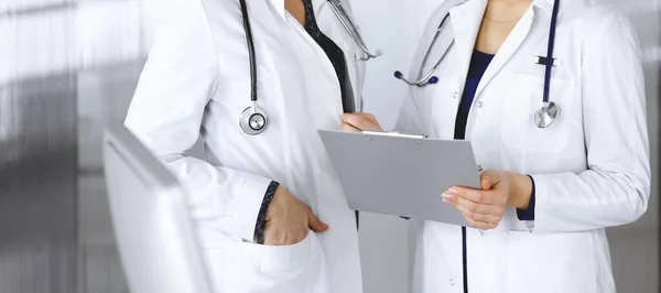 Δύο γυναίκες γιατροί συζητούν για τις ιατρικές εξετάσεις των ασθενών τους, ενώ στέκονται σε ένα γραφείο κλινικής. Οι γιατροί χρησιμοποιούν ένα πρόχειρο στη δουλειά. Ομαδική εργασία στην ιατρική — Φωτογραφία Αρχείου