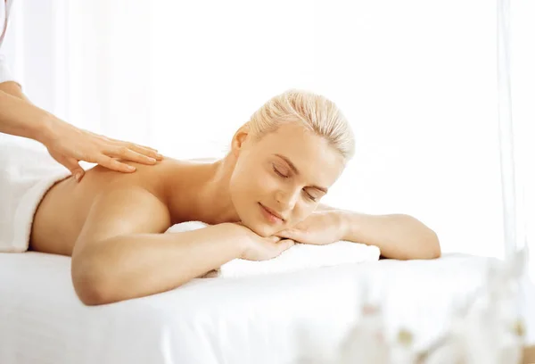 Beautiful blonde woman enjoying back massage with closed eyes in sunny spa salon. Beauty concept