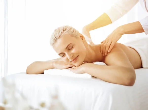 Beautiful blonde woman enjoying back massage with closed eyes in sunny spa salon. Beauty concept