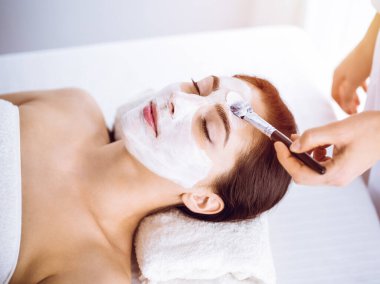 Güzel esmer kadın güneşli spa merkezinde kapalı gözlerle kozmetik maske takmaktan zevk alıyor. Rahatlatıcı tedavi ve ilaç konsepti