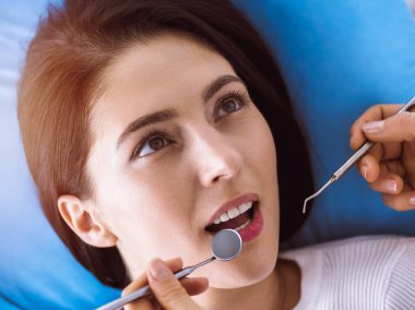 Güneşli diş kliniğinde dişçi tarafından muayene edilen gülümseyen esmer kadın. Hastanın ağzının yanında diş aletleri tutan bir doktorun elleri. Sağlıklı diş ve ilaç konsepti