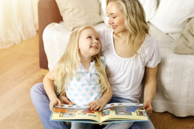 Mutlu bir aile. Sarışın genç anne güneşli odada ahşap zeminde otururken sevimli kızına kitap okuyor. Annelik kavramı