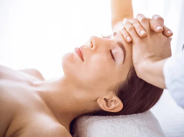 Piękna brunetka korzystająca z masażu twarzy z zamkniętymi oczami w słonecznym centrum spa. Relaksujące koncepcje leczenia i medycyny kosmetycznej — Zdjęcie stockowe