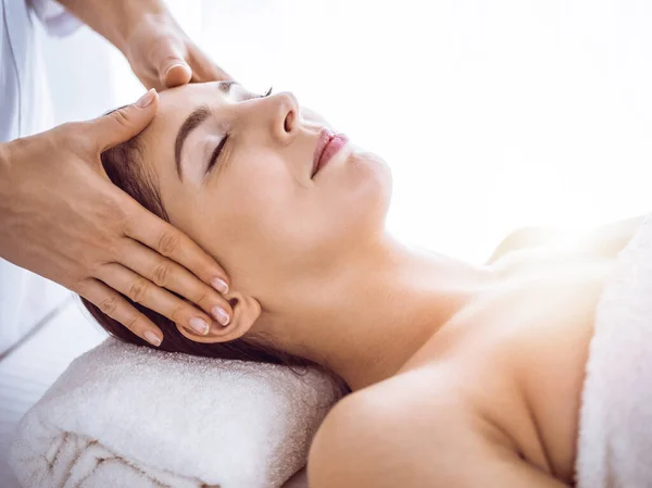 Hermosa mujer morena disfrutando de masaje facial con los ojos cerrados en el soleado centro de spa. Tratamiento relajante y conceptos de medicina cosmética — Foto de Stock