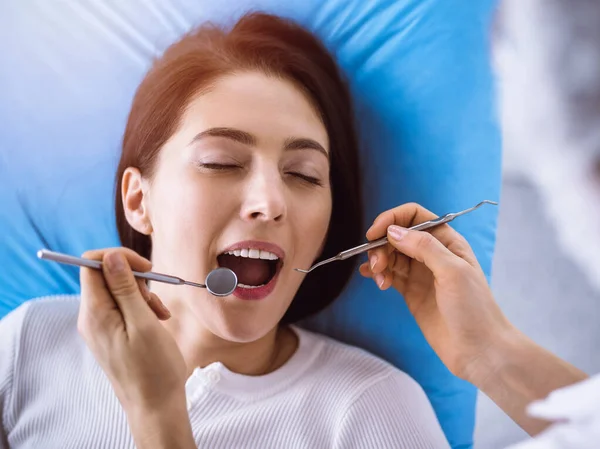 Uśmiechnięta brunetka badana przez dentystę w słonecznej klinice dentystycznej. Ręce lekarza trzymającego instrumenty stomatologiczne w pobliżu ust pacjenta. Zdrowe zęby i koncepcja medycyny — Zdjęcie stockowe