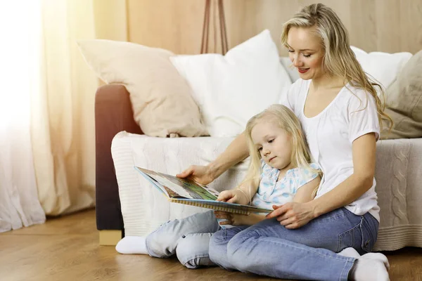 Bonne famille. Jeune mère blonde lisant un livre à sa jolie fille assise au sol en bois dans une pièce ensoleillée. Concept de maternité — Photo