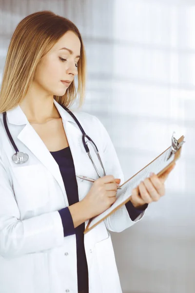 Jeune femme-médecin intelligente prescrit des médicaments pour un patient, à l'aide d'un presse-papiers, tout en se tenant dans l'armoire dans une clinique. Médecine avec stéthoscope sur son lieu de travail — Photo