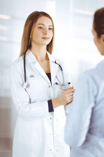 Jovem médica e sua paciente estão discutindo os pacientes exame de saúde atual, enquanto estão juntos em um consultório hospitalar. A médica está a escrever algumas marcas, usando uma prancheta. Perfeito. — Fotografia de Stock