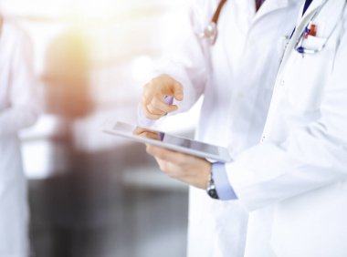 Bilinmeyen bir grup doktor, güneşli bir hastane ofisinde dururken bazı tıbbi isim kayıtlarını kontrol etmek için bir bilgisayar tableti kullanıyor. Doktorlar hastaları muayene etmeye ve yardım etmeye hazır. Tıbbi yardım, sigorta.