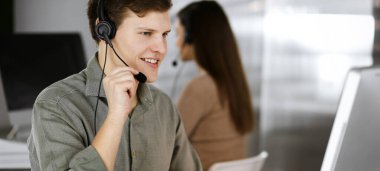 Yeşil tişörtlü ve kulaklıklı genç siyah saçlı bir adam, masada oturmuş, modern bir ofiste bayan bir meslektaşıyla birlikte çalışırken, bir müşteriyle konuşuyor. İş yerindeki çağrı merkezi operatörleri