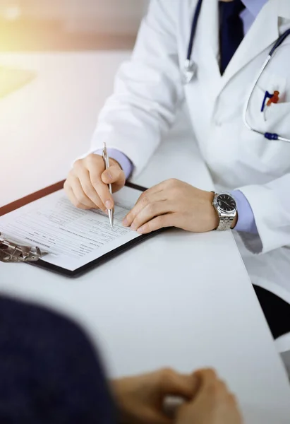 Un médico está escribiendo una receta médica para su paciente, mientras está sentado junto al escritorio en el gabinete soleado de un hospital. Médico que usa portapapeles para llenar los registros del historial de medicamentos — Foto de Stock