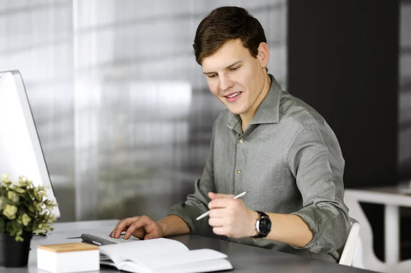 Jeune homme d'affaires brun et programmeur dans une chemise verte écrit quelques notes dans son plan d'affaires, tout en étant assis sur le bureau dans un cabinet moderne. Prise de vue ou portrait d'entreprise dans un bureau — Photo