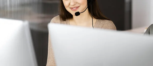 Jeune fille amicale dans les écouteurs parle à un client des entreprises, tout en étant assis sur le bureau dans un bureau moderne avec son collègue. Opérateurs de centre d'appels au travail — Photo