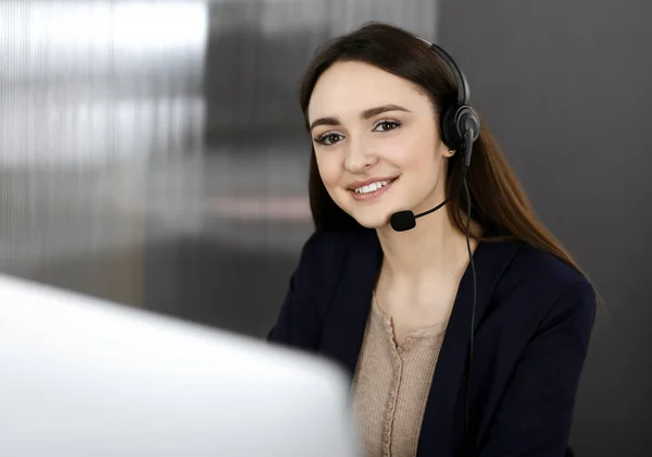 Joven chica sonriente en auriculares está hablando con el cliente de las empresas, mientras está sentado en el escritorio en una oficina de la compañía. Retrato de un operador de centro de llamadas en el trabajo — Foto de Stock