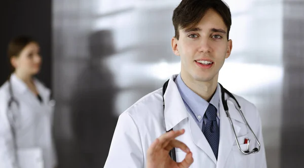 Доброзичливий чоловік-лікар стоїть прямо і показує знак ОК в клініці. Жінка-колега поспішає на тлі лікаря. Концепція медицини — стокове фото