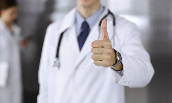 Φιλικός άνθρωπος-γιατρός στέκεται ευθεία και δείχνει Ok υπογράψει με τους αντίχειρες επάνω στη σύγχρονη κλινική. Γυναίκα συνάδελφος είναι σε μια βιασύνη στο παρασκήνιο του γιατρού. Έννοια ιατρικής — Φωτογραφία Αρχείου