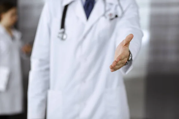 Unknown man-doctor standing straight and offering helping hand in modern clinic. Female colleague is in a hurry at the background of physician. Medicine concept