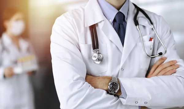 Unknown male doctor in protective mask standing with arms crossed in sunny clinic. Medicine concept during Coronavirus pandemic — Stock Photo, Image