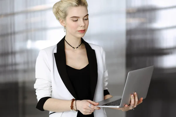 Young blonde business woman or female student in white suit is standing in modern office. Lifestyle and diverse people concept