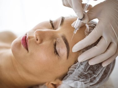 Güzellik uzmanı güneşli spa merkezinde genç esmer bir kadının yüzüne şırıngayla güzellik prosedürü uyguluyor. Kozmetik ilaç ve ameliyat, güzellik iğneleri.