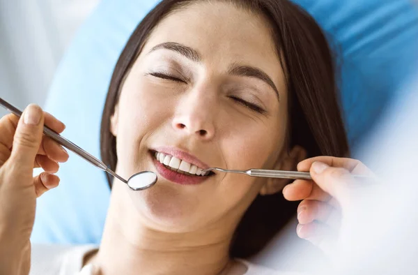 Lächelnde brünette Frau, die vom Zahnarzt in der Zahnklinik untersucht wird. Die Hände eines Arztes halten zahnärztliche Instrumente in der Nähe des Mundes des Patienten. Gesunde Zähne und medizinisches Konzept — Stockfoto