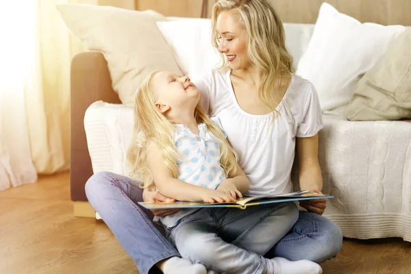 Happy family. Blonde young mother reading a book to her cute daughter while sitting at wooden floor in sunny room. Motherhood concept