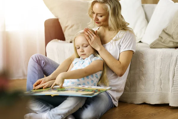 Glückliche Familie. Blonde junge Mutter liest ihrer süßen Tochter ein Buch vor, während sie am Holzboden in einem sonnigen Raum sitzt. Mutterschaftskonzept — Stockfoto