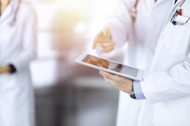 Bilinmeyen bir grup doktor, güneşli bir hastane ofisinde dururken bazı tıbbi isim kayıtlarını kontrol etmek için bir bilgisayar tableti kullanıyor. Doktorlar hastaları muayene etmeye ve yardım etmeye hazır. Tıbbi yardım, sigorta.