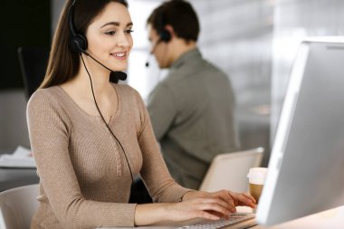 Modern bir ofiste meslektaşıyla birlikte masa başında otururken, kulaklıklı genç bir kız bir firma müşterisiyle konuşuyor. İş yerindeki çağrı merkezi operatörleri