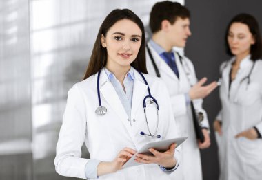 Gülümseyen zeki kadın-doktor, klinikte meslektaşlarıyla birlikte ayakta dururken elinde bir tablet bilgisayar tutuyor. Doktorlar iş başında. Hastanede mükemmel bir tıbbi hizmet.