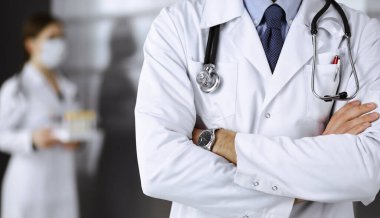 Koruyucu maskeli kimliği belirsiz erkek doktor klinikte kollarını kavuşturmuş duruyor. Coronavirus salgını sırasında ilaç konsepti. Covid 2019