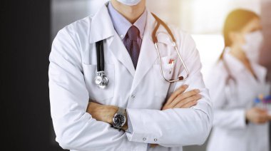 Güneşli klinikte kolları çapraz duran koruyucu maskeli kimliği belirsiz erkek doktor. Coronavirüs salgını sırasında ilaç kavramı