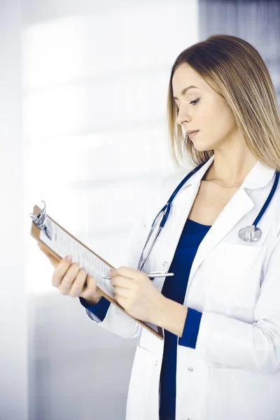 Jovem mulher-médico inteligente está prescrevendo algum medicamento para um paciente, usando uma área de transferência, enquanto está no gabinete em uma clínica. Médica com estetoscópio no local de trabalho — Fotografia de Stock