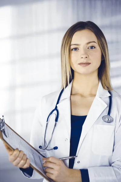 Молода розумна жінка-лікар призначає пацієнту ліки, використовуючи буфер обміну, стоячи в кабінеті в клініці. Жінка-лікар зі стетоскопом на робочому місці — стокове фото
