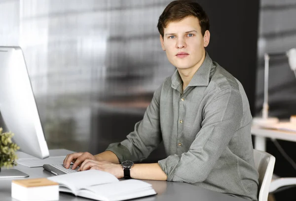 Jonge succesvolle zakenman en programmeur in een groen shirt kijkt naar de camera, terwijl hij aan zijn computer werkt, zittend achter het bureau in een kast. Hoofdfoto of bedrijfsportret in een kantoor — Stockfoto