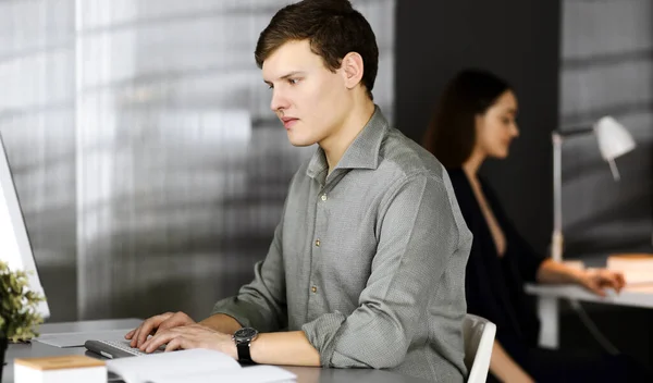Un jeune homme d'affaires travailleur en chemise verte est à la recherche d'informations sur son ordinateur, assis au bureau dans un cabinet moderne avec une collègue féminine en arrière-plan. Tête de mort ou — Photo