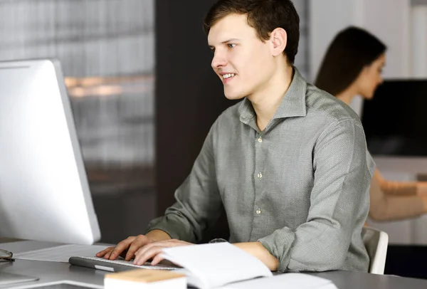Темноволосый молодой бизнесмен и программист в зеленой рубашке усердно работает за компьютером, сидя за столом в современном кабинете с коллегой-женщиной на заднем плане. Концепция — стоковое фото