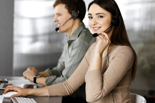 Kulaklık takmış iki genç bir ofiste masada otururken müşterilerle konuşuyor. Kadınlara odaklan. İş yerindeki çağrı merkezi operatörleri — Stok fotoğraf