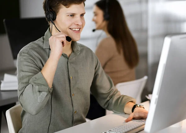 Young dark-haired guy in a green shirt and headsets is talking to a client, while sitting at the desk, working together with a female colleague in a modern office. Call center operators at work — Stock Photo, Image
