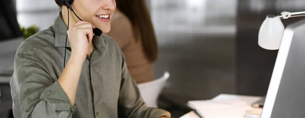 Un jeune homme aux cheveux bruns portant une chemise verte et des écouteurs parle à un client, assis au bureau, travaillant avec une collègue dans un bureau moderne. Opérateurs de centre d'appels au travail — Photo